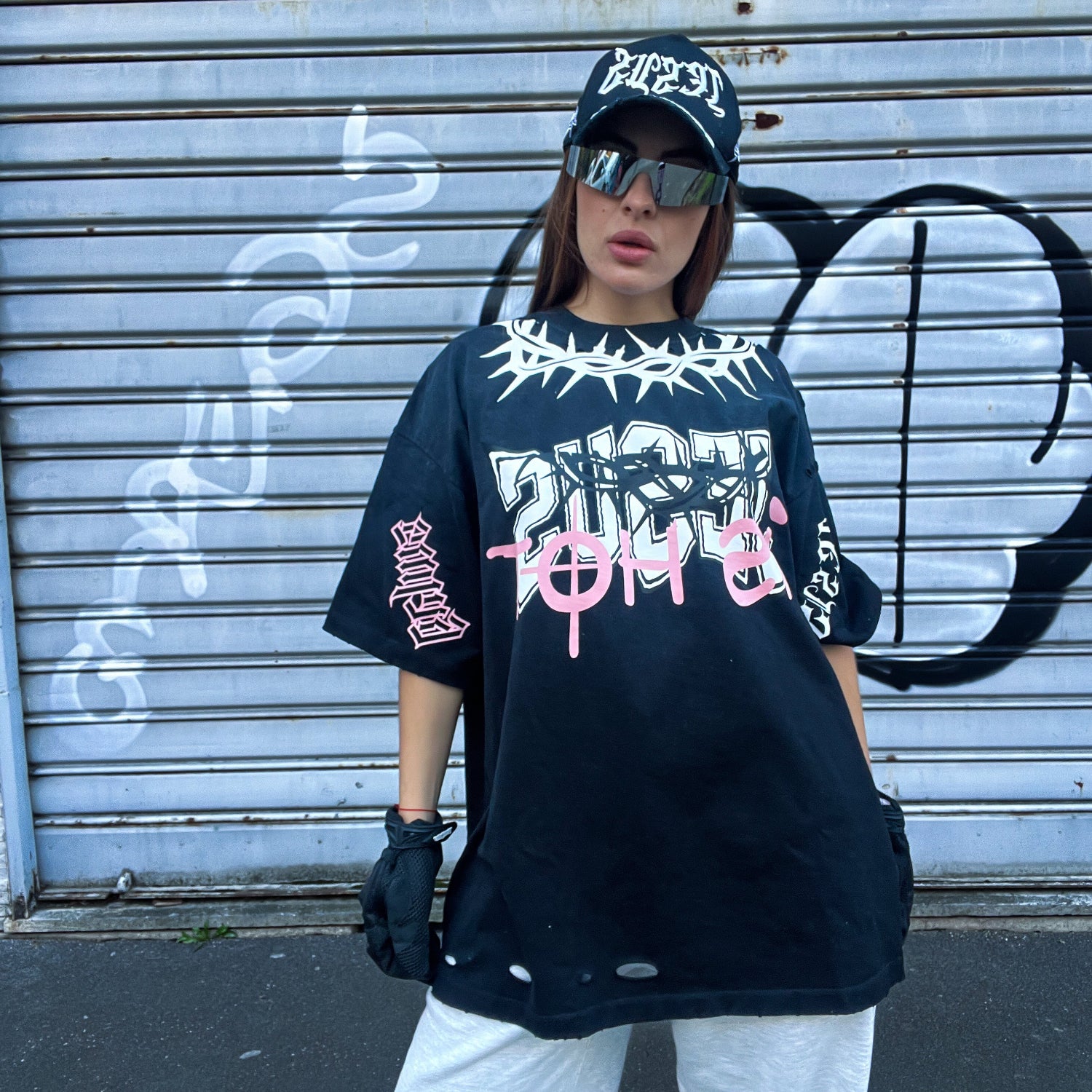 Person wearing a black oversized t-shirt with graphic designs, white pants, and a cap in front of a graffiti-covered wall.