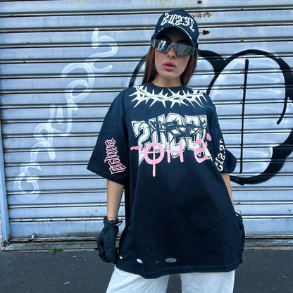 Person wearing a black oversized t-shirt with graphic designs, white pants, and a cap in front of a graffiti-covered wall.